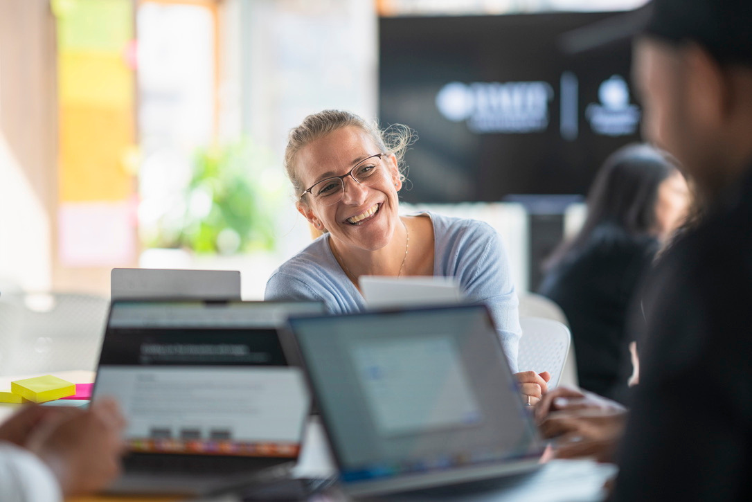 Picture of Steph smiling. 3 MacBook in the foreground. 
Steph is wearing glasses and that same light blue top. 